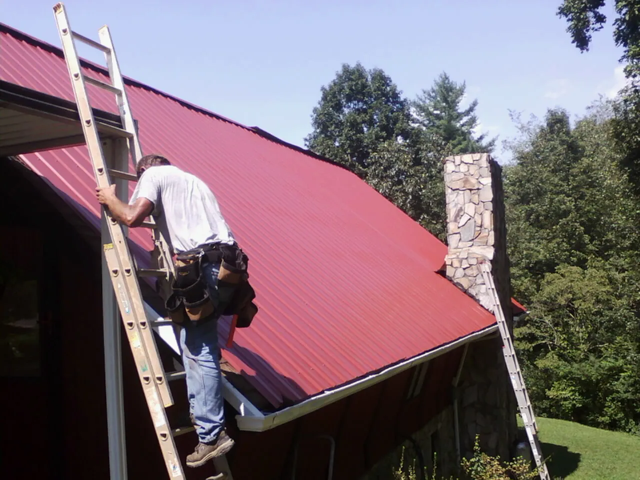 Roofer on a ladder beside a red metal roof and stone chimney — Soffit & Fascia Repair by Roofing Brothers in Dighton